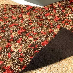 May include: A rectangular fabric with a pattern of brown pinecones and red berries on a dark brown background. A section of black, textured fabric is visible on the right side, suggesting a reversible design or contrasting element.