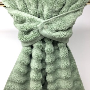 May include: A sage green kitchen towel with a textured waffle pattern and a button closure. The towel is hanging on a silver towel rack.