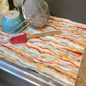 May include: A colorful striped kitchen dish drying mat with a red spatula, a wooden spoon, and a silver strainer on top.
