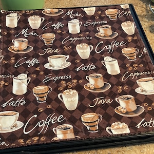 May include: A brown quilted fabric hot pad with a coffee theme. The pad is decorated with various coffee cups, coffee beans, and the words "Mocha", "Caffe", "Java", "Expresso", "Cappuccino", "Latte", and "Coffee".