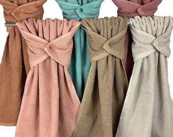 May include: A collection of six hanging kitchen towels in various solid colors including terracotta, teal, dusty rose, tan, and cream. Each towel features a textured fabric and a top flap secured with a small matching plastic snap button.
