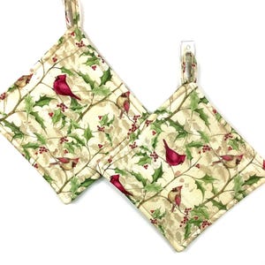 May include: Two square quilted pot holders with a cream background featuring a pattern of red cardinals, green holly leaves, and red berries. Each pot holder has a loop for hanging.