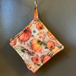 May include: A square, quilted pot holder featuring a fall harvest theme. The fabric is patterned with orange, white, and green pumpkins, along with autumn leaves and berries. The pot holder has a loop for hanging.