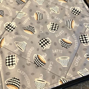 May include: A gray quilted fabric with a coffee-themed pattern. The design features various coffee cups in black and white, along with coffee beans and text like "Cappuccino", "Espresso", and "Latte". The fabric is bordered with a black trim.