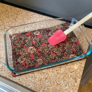 May include: A clear glass baking dish with a red silicone spatula and a patterned fabric liner featuring pinecones and red berries. The spatula has a wooden handle. The dish is on a speckled countertop.