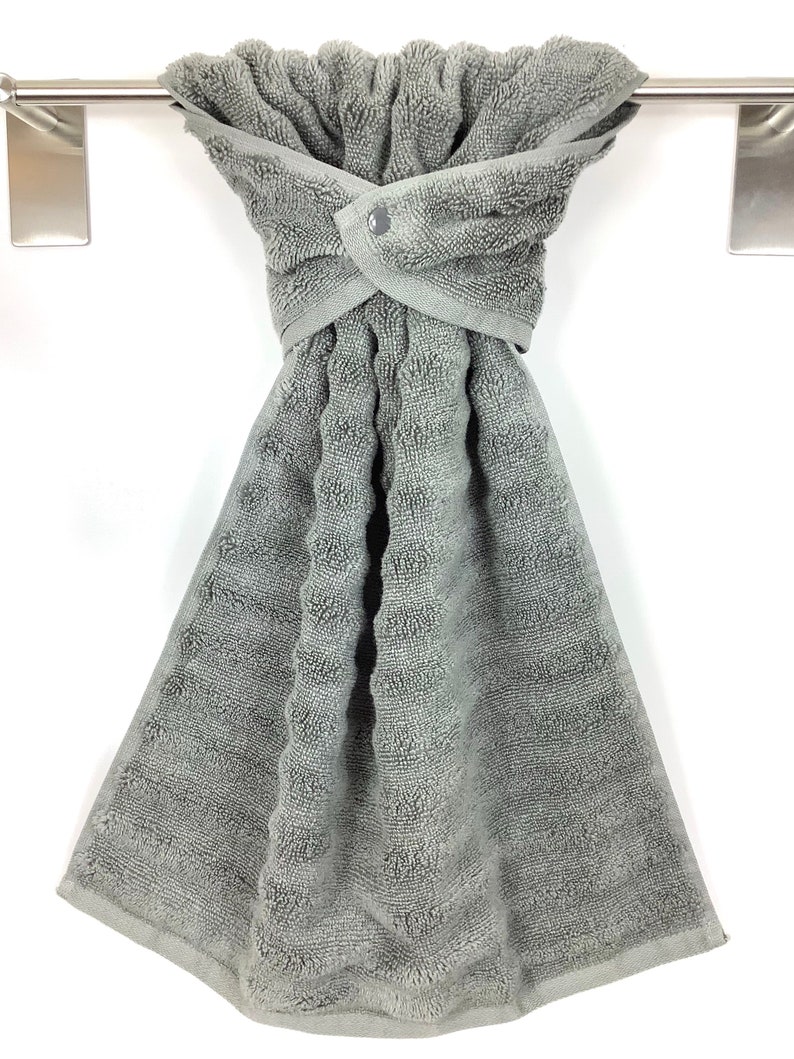 May include: A gray kitchen towel with a button closure hangs on a silver towel rack. The towel is made of a soft, absorbent material and is perfect for drying dishes or hands.