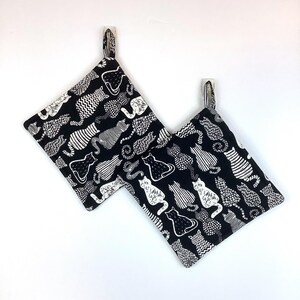 May include: Two black and white cat-themed pot holders. The pot holders are square-shaped with a loop for hanging. The fabric features a repeating pattern of various cat illustrations in white against a black background. The pot holders are hanging on a white wall.