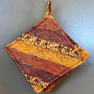 May include: A square quilted potholder featuring a diagonal striped pattern in shades of deep burgundy, burnt orange, and golden yellow. The fabric has a textured, woven appearance with a small fabric loop at the top corner for hanging purposes.