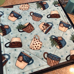 May include: A blue and white quilted hot pad with a teacup pattern. The teacups are brown, blue, and white with polka dots and floral designs. The hot pad has a black border.