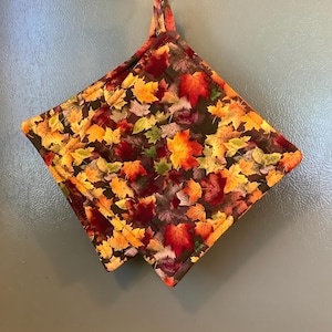 May include: A square quilted potholder featuring a dark brown background with a dense pattern of autumn leaves in shades of red, orange, yellow, and green. The potholder has a fabric loop at the top corner for hanging and is stitched with a border.