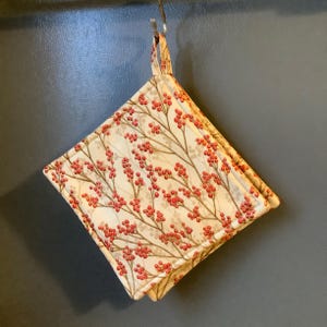 May include: A square, quilted pot holder with a loop for hanging. The fabric features a cream background with a pattern of red berries on brown branches. The pot holder is hanging from a metal hook.
