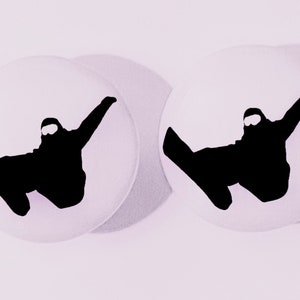 May include: Two round, white buttons with a black silhouette of a snowboarder in mid-air. The snowboarder is wearing goggles and a helmet, with arms outstretched. The buttons have a shadow effect.