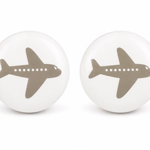 May include: Two round white cabinet knobs, each featuring a centered taupe-colored airplane graphic. The airplane design includes a series of small white dots along the fuselage to represent windows, set against the smooth, glossy white surface.