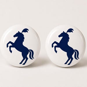 May include: Two circular white ceramic knobs, each featuring a dark navy blue silhouette of a prancing horse with a plume on its head. The design is centered on the smooth, glossy surface of each knob, providing a clear and consistent graphic.