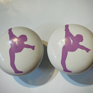 May include: Two white ceramic knobs with a purple silhouette of a person in a yoga pose.