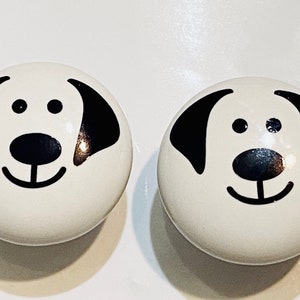 May include: Two white ceramic knobs with black dog faces. The dog faces have black eyes, noses, and mouths.