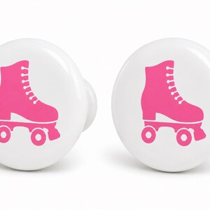 May include: Two round white ceramic cabinet knobs, each featuring a bright pink graphic of a quad roller skate centered on the front surface. The knobs have a smooth, glossy finish and a simple, circular shape with a protruding base for mounting.