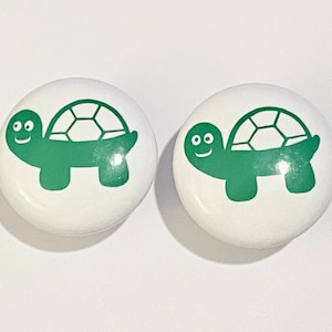 May include: Two white ceramic knobs with a green turtle design. The turtles have a smiling face and a shell with a pattern.