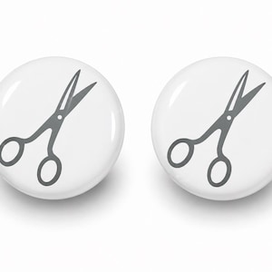 May include: Two circular white pin-back buttons, each featuring a centered dark gray graphic illustration of a pair of open scissors. The buttons have a smooth, glossy finish and a simple, clean design with a clear, high-contrast icon.