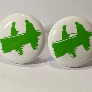May include: Two white ceramic buttons with a green silhouette of two people in a boat. The silhouette is stylized and abstract.