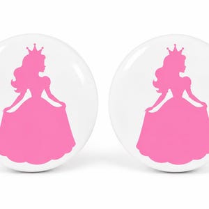 May include: Two round white cabinet knobs, each featuring a bright pink silhouette of a princess wearing a crown and a long gown. The design is centered on the smooth, glossy surface of the circular knobs, which have a simple, clean aesthetic.