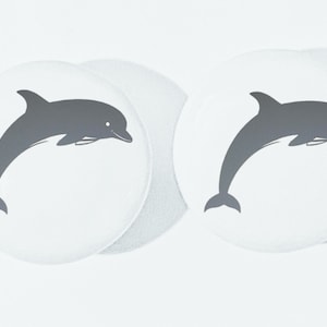 May include: Two white round coasters with a gray silhouette of a dolphin jumping out of the water on each coaster.