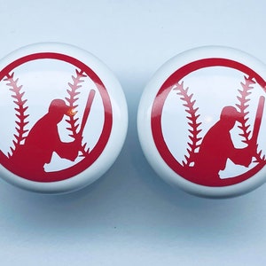 May include: Two white cabinet knobs with a red baseball design. Each knob features a red silhouette of a baseball player with a bat, set against a baseball's stitching. These knobs are perfect for a sports-themed room.