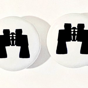 May include: Two circular white buttons, each featuring a bold, solid black silhouette of a pair of binoculars centered on the surface. The design is simple and high-contrast, displaying the distinct shape of the optical instrument clearly.