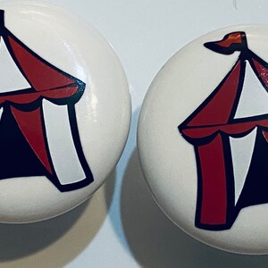 May include: Two white ceramic cabinet knobs with a red and white circus tent design. The tent has a black outline and a red flag on top.