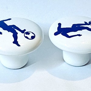 May include: Two white ceramic cabinet knobs with a blue silhouette of a soccer player kicking a ball.