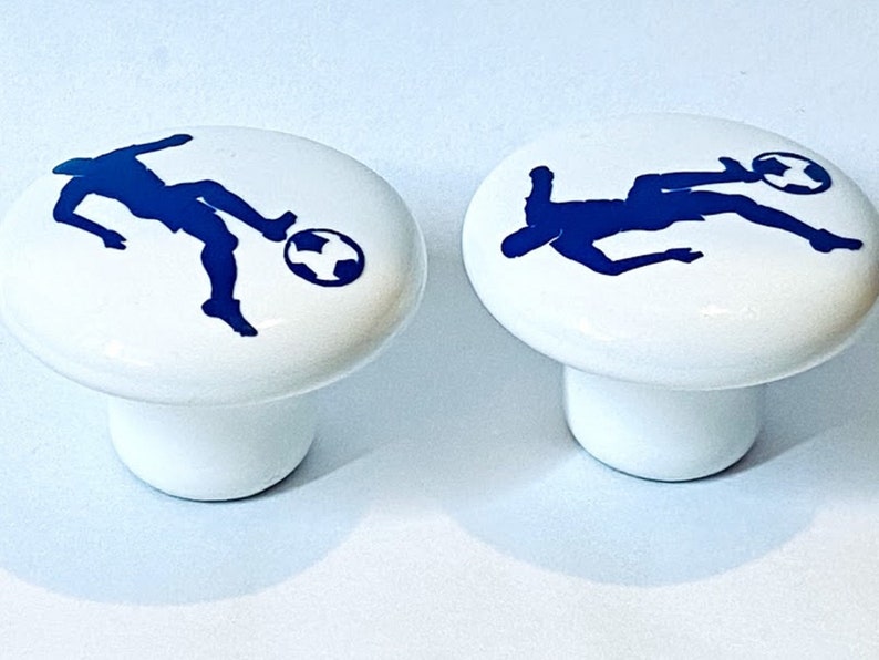 May include: Two white ceramic cabinet knobs with a blue silhouette of a soccer player kicking a ball.