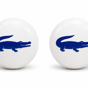May include: Two spherical white ceramic knobs, each featuring a centered, solid dark blue crocodile graphic. The smooth, glossy finish of the round knobs highlights the contrast between the white surface and the bold blue animal silhouette.