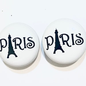 May include: Two white round knobs with a black silhouette of the Eiffel Tower and the word "PARIS" in a decorative font.