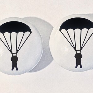 May include: Two round white ceramic knobs, each featuring a black graphic print of a person descending with an open parachute. The knobs have a smooth, glossy finish and a domed shape, displaying a clear silhouette of a skydiver in mid-air.