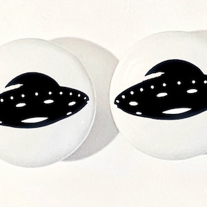 May include: Two white round buttons with a black silhouette of a UFO on each. The UFOs have a rounded top and a flat bottom with a row of small circles along the bottom edge.