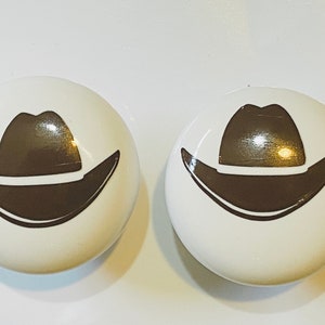 May include: Two white ceramic knobs with a brown cowboy hat design.