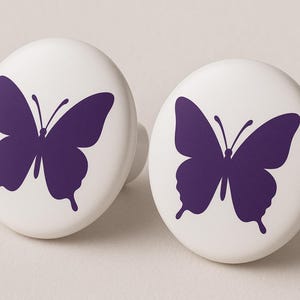May include: Two white cabinet knobs with a purple butterfly design. The butterfly is centered on the round knob. The knobs are made of ceramic or porcelain and have a small, rounded base.