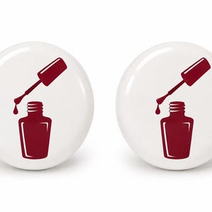 May include: Two circular white buttons, each featuring a dark red graphic illustration of an open nail polish bottle with a brush applicator hovering above it, showing a single drop of polish falling from the tip of the brush into the bottle.