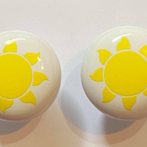 May include: Two white ceramic knobs with a yellow sun design. The sun has a yellow center and yellow rays.