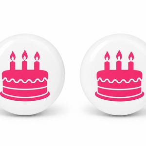 May include: Two circular white buttons, each featuring a vibrant pink graphic of a birthday cake topped with three lit candles. The cake design includes a scalloped frosting detail along the middle and a solid base, presented in a clean, flat style.