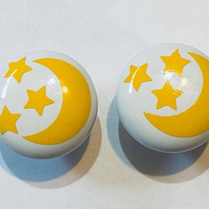 May include: Two white ceramic knobs with a yellow crescent moon and three yellow stars design.