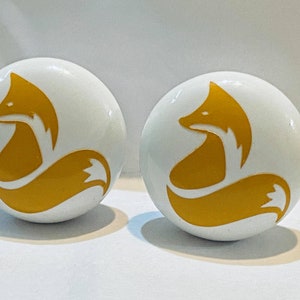 May include: Two white ceramic knobs with a gold fox silhouette design. The fox is curled up with its tail wrapped around its body.