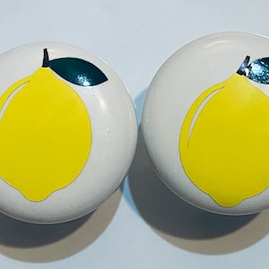 May include: Two white ceramic knobs with a yellow lemon design. Each knob features a green leaf and a black stem.