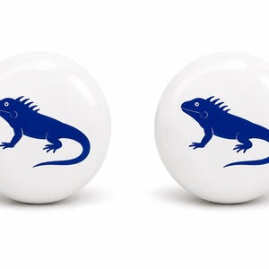 May include: Two round, white ceramic knobs, each featuring a centered, dark blue graphic illustration of an iguana in profile. The knobs have a smooth, glossy finish and a spherical shape, providing a clean, uniform appearance for furniture hardware.