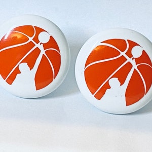 May include: Two white round ceramic buttons with an orange basketball and a white silhouette of a person shooting a basketball.