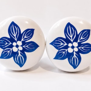 May include: Two round white ceramic cabinet knobs, each featuring a hand-painted blue floral design with six pointed petals and a central cluster of four small white dots. The knobs have a smooth, glossy finish and a circular, convex shape.