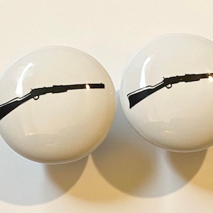 May include: Two white ceramic knobs with a black silhouette of a rifle printed on each.