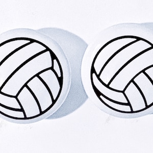 May include: Two white round buttons with a black and white volleyball design. The volleyball graphic features black lines outlining the panels of the ball. The buttons are set against a white background.