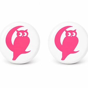 May include: Two circular white buttons featuring a vibrant pink graphic of an owl perched on a crescent moon. The design is centered on each button, showcasing a bold contrast between the bright pink illustration and the solid white background.