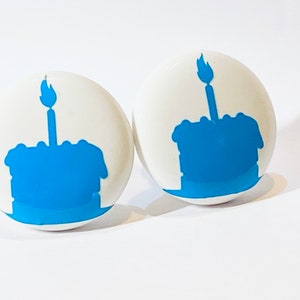 May include: Two white round ceramic buttons with a blue silhouette of a birthday cake with a single candle on top.